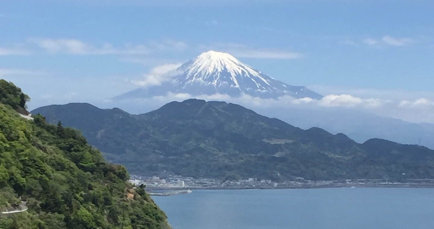 Snow-capped Mount Fuji and Suruga Bay seen from Satta Pass, with hills in front of Fuji, a town along the shore and a green slope in the foreground on the left